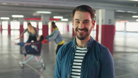 portrait of caucasian man standing in front of the camera and smiling while on the background his friends having fun with trolley in the parking