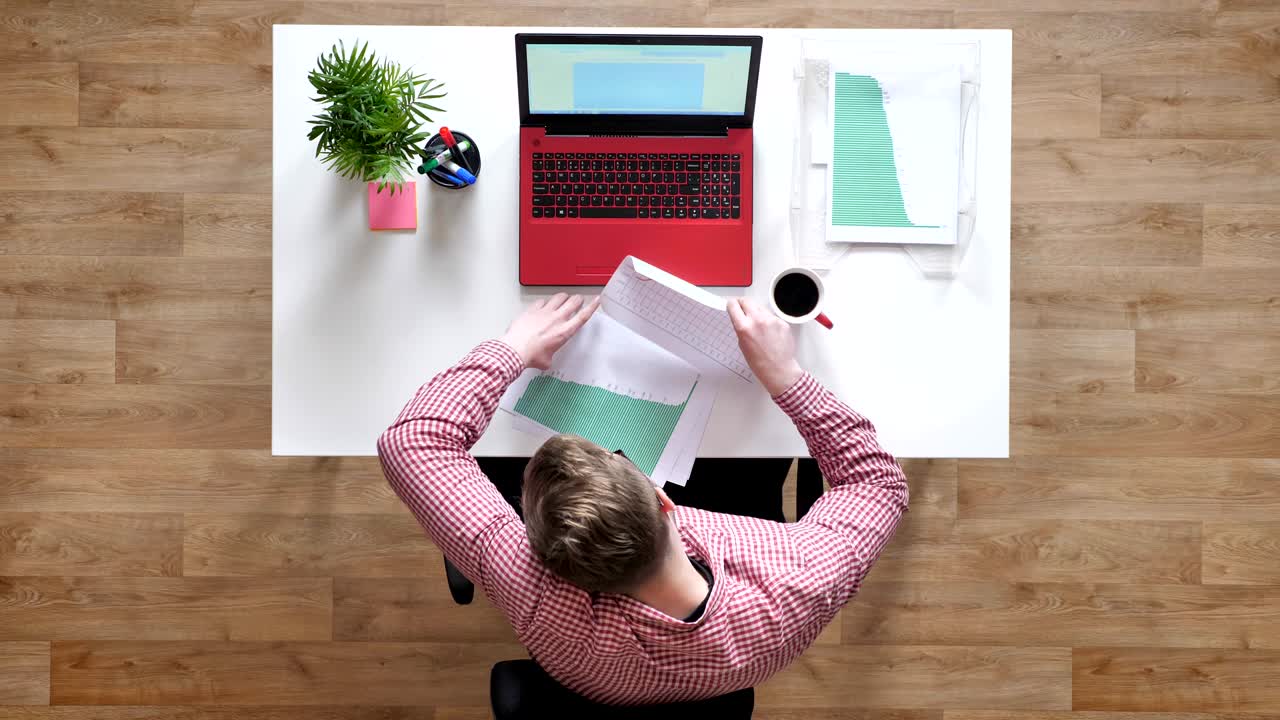 joven con gafas escribiendo en la computadora portátil, hablando por teléfono y tomando documentos, ocupado, topshot, sentado detrás de la mesa