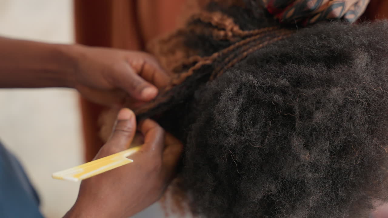 Primo piano delle mani che separano i capelli naturali, stilista che separa riccioli stretti con un pettine a denti fini preparando trecce cornrow ordinate, scena intima di salone casalingo con movimenti lenti e deliberati, tensione delicata e concentrata