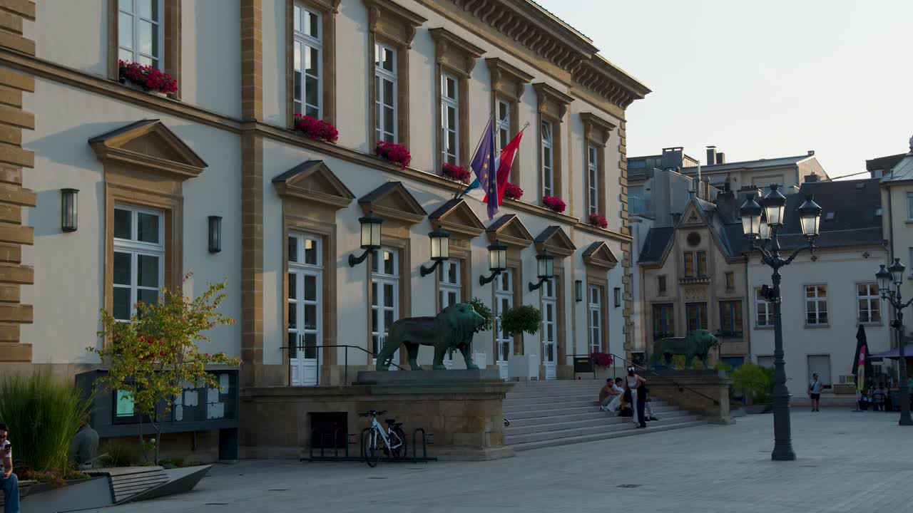 Static wide shot of city hall, lion statue, flags, and people in soft evening light