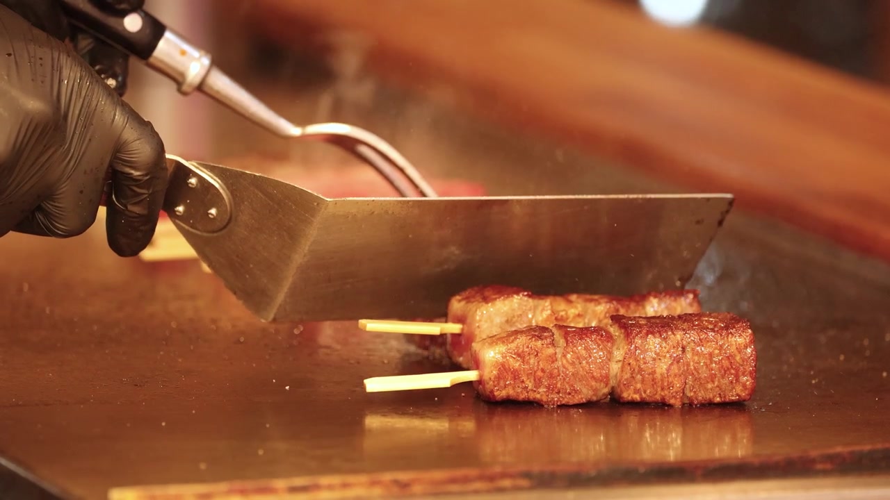 Primo piano di uno chef che usa una spatola per premere spiedini sfrigolanti di manzo wagyu su una griglia calda.