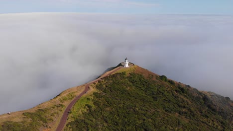 Aerial-view-of-the-iconic-Cape-Reinga-Lighthouse-on-the-north-edge-of-New-Zealand-on-early-sunny-morning,-standing-above-clouds-in-4K
