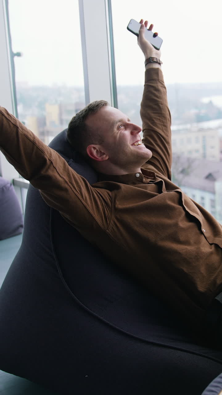 Relaxed businessman indoors. Happy young man is sitting by the window with stretched arms and having a rest in office on the city view window background. Vertical video