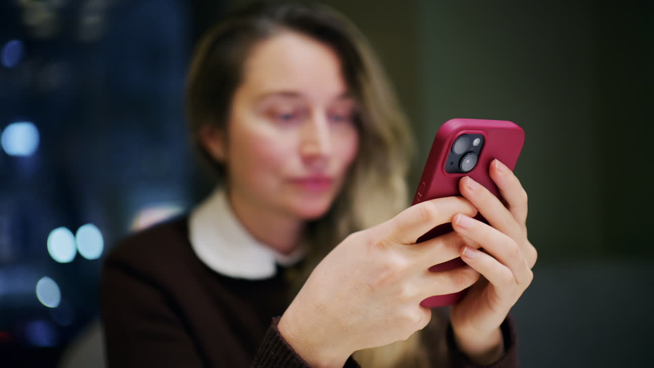 Una donna in casa di notte, illuminata da una luce soffusa, guarda il suo telefono con un leggero sorriso.