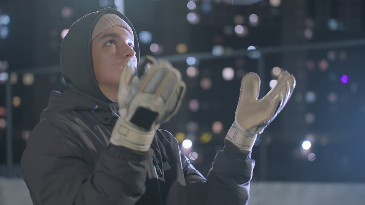 장갑을 입은 남자가 축구공을 공중으로 던지고 밤에 강렬한 훈련 중에 잡는 클로즈업, 흐릿한 배경에는 높은 건물과 반이는 도시 불빛이 있습니다.