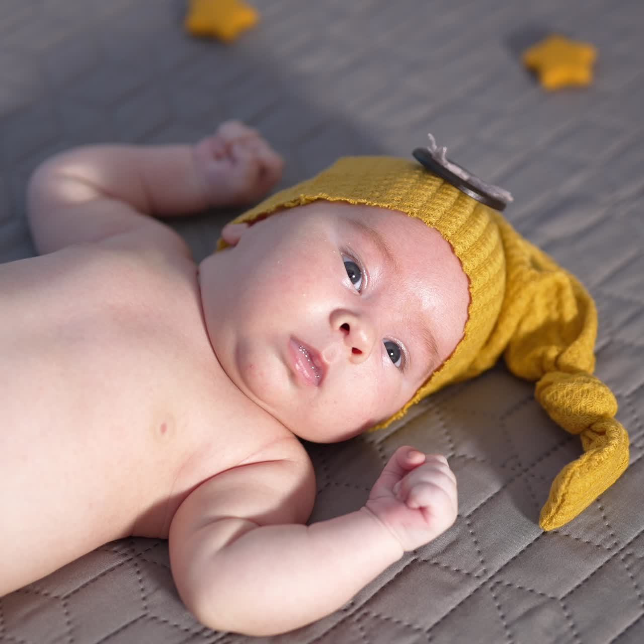 Infant boy with naked belly lying still on the bed. Baby in a yellow cap and pants looks intensely into the camera. Close up