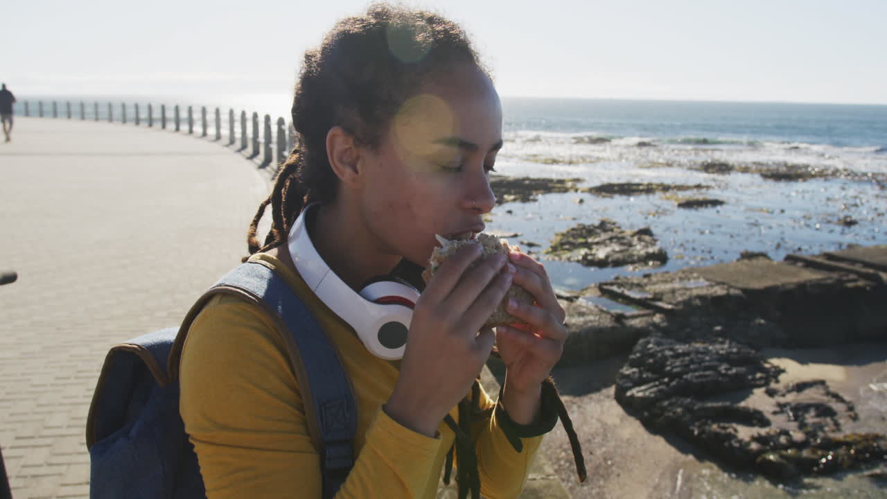 African american woman sitting eating sandwich promenade by the sea