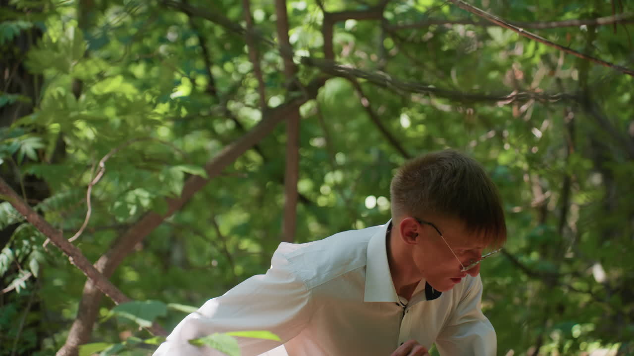 Close view of man in white shirt carrying backpack bending carefully under branches while walking through dense green forest in daylight, moving cautiously with focus on exploration