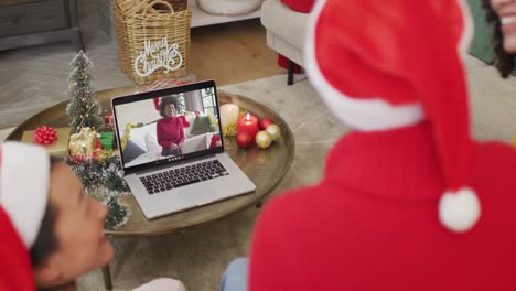 Familia-Diversa-Con-Sombreros-De-Santa-Usando-Una-Computadora-Portátil-Para-Una-Videollamada-Navideña-Con-Una-Mujer-Feliz-En-La-Pantalla