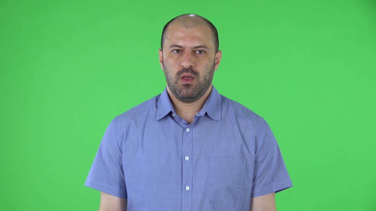 Portrait of a middle aged men looking at the camera in anticipation and then very upset. Balding male with beard in blue shirt posing on green screen in the studio. Close up
