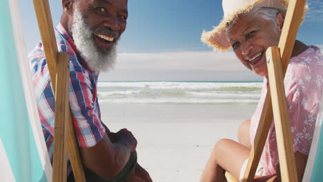 una pareja de mayores de raza mixta sentada en camas de sol y sonriendo en la playa