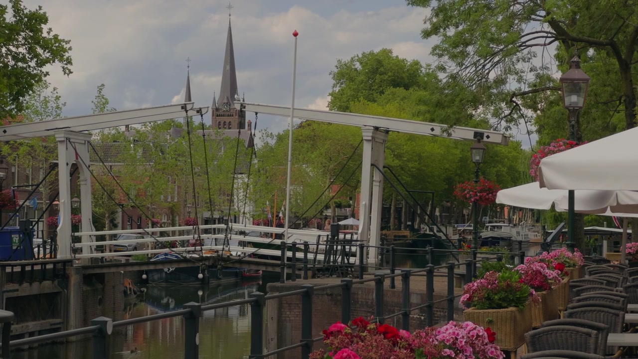Typische holländische Postkarten-Stadtszene mit einer Kanalbrücke, einem historischen Kirchturm, Café-Terrassen mit Sonnenschirmen, bunten Blumen, Laubbäumen und einem klaren blauen Sommerhimmel