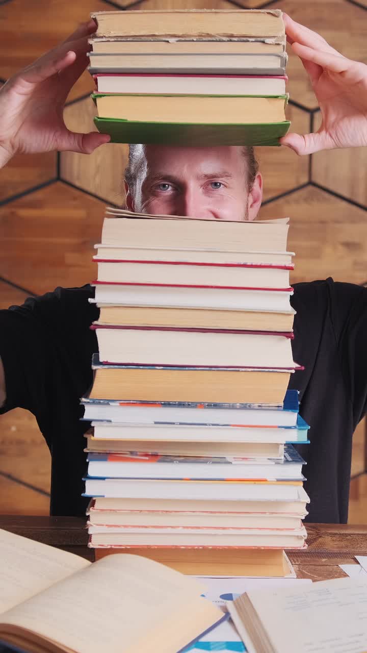 Young positive caucasian man with stack of books on table sits in library