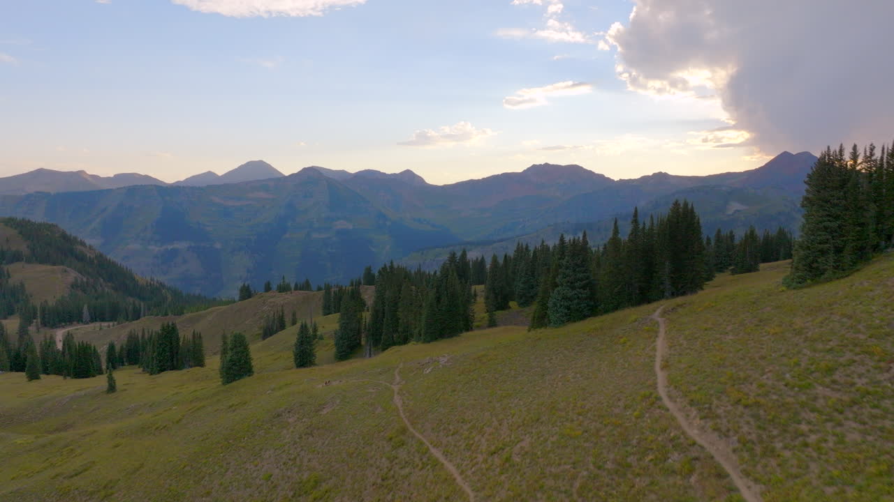 Ripresa aerea che sorvola un sentiero escursionistico e una cresta alberata e si dirige verso una catena montuosa nelle Montagne Rocciose del Colorado al tramonto