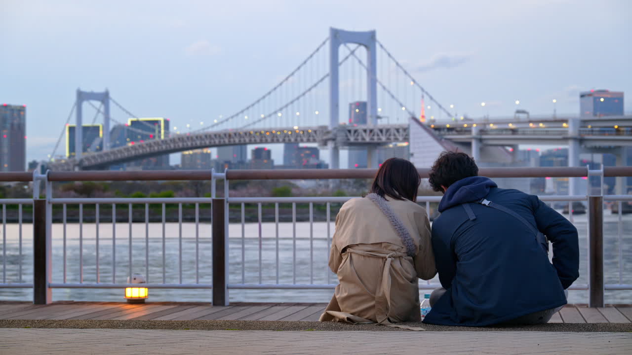 A man and a woman couple sitting in front of a view of the Rainbow Bridge and the skyline of Tokyo, Japan in the evening