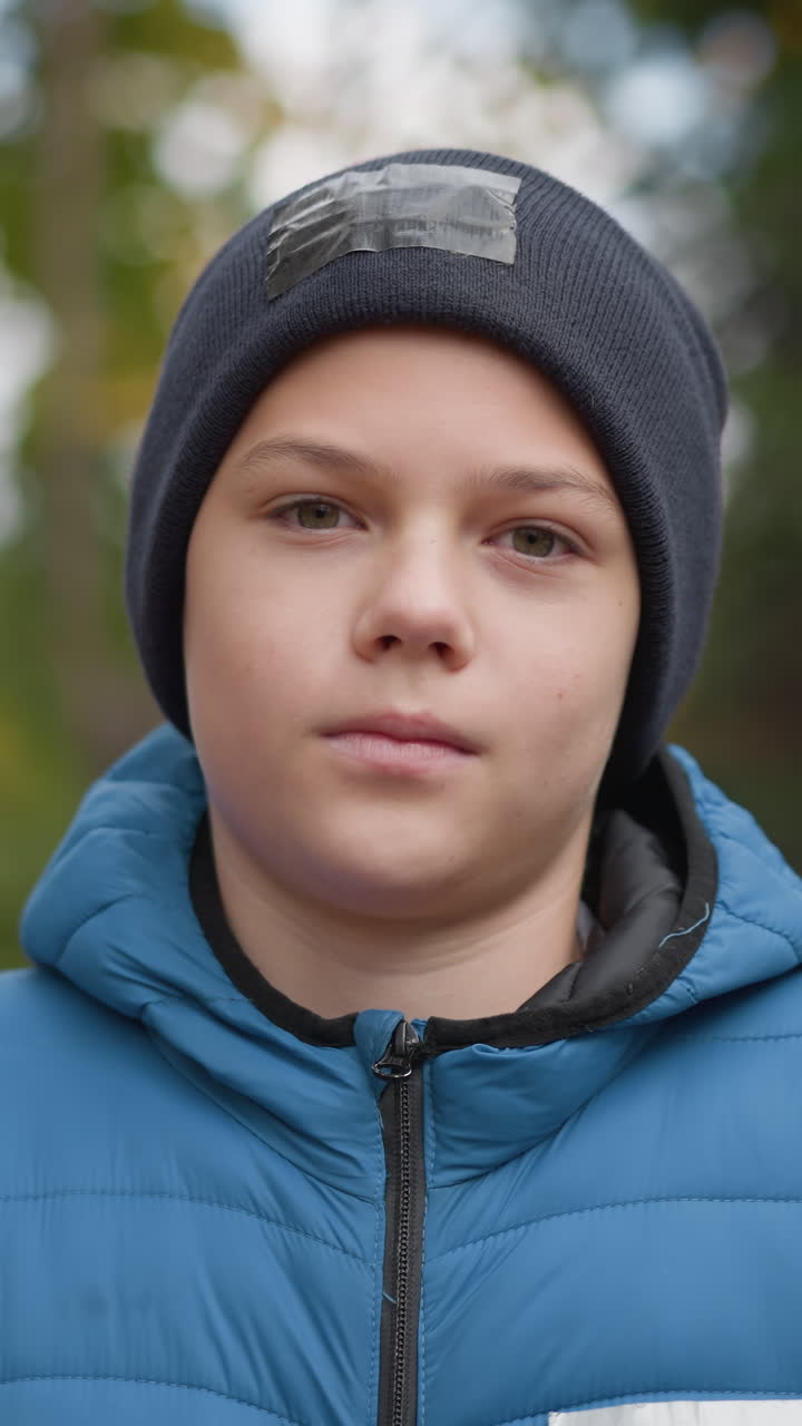 niño con gorra negra y chaqueta azul de pie al aire libre, ajustando y cerrando su chaqueta en un día fresco de otoño, rodeado de árboles borrosos y naturaleza