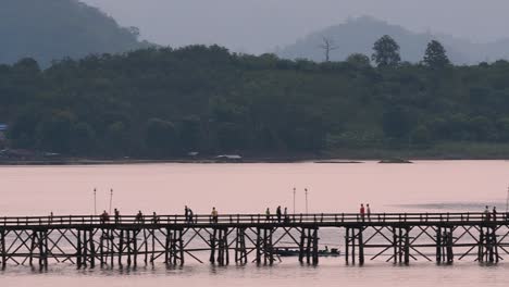 Mon-Bridge-and-a-longboat-followed-speeding-behind-the-structure-while-people-are-walking-on-the-bridge,-silhouetting-as-it-was-getting-dark,-in-slow-motion