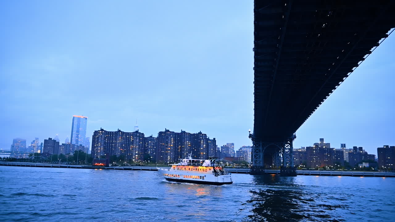 Riverboat moves under the bridge at dusk time. River tour by New York, USA after sunset. City skyline at backdrop