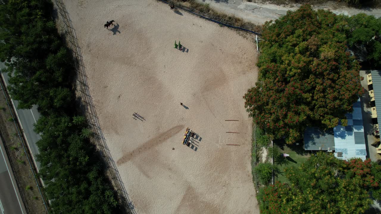 Top down ascending aerial reveals stables and two riders riding an obstacle course inside the equestrian center