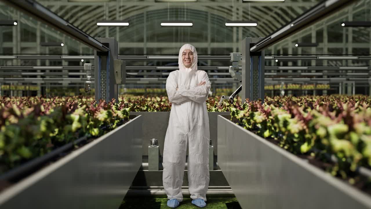 cuerpo lleno de hombre asiático investigador sonriendo y cruzando los brazos mientras estaba de pie en el invernadero con agricultores robóticos inteligentes