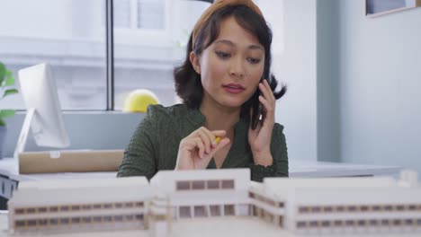 Biracial-businesswoman-talking-on-smartphone-and-building-model-alone-at-office