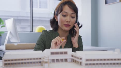 mujer de negocios biracial hablando en un teléfono inteligente y construyendo un modelo sola en la oficina