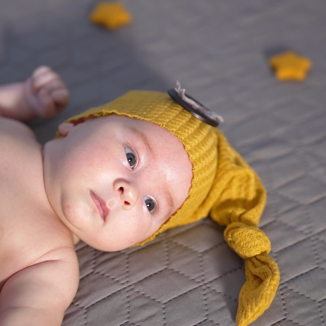 Bare baby in a funny yellow cap lying on the bed. Small boy lying calmly on the bed and look at the camera
