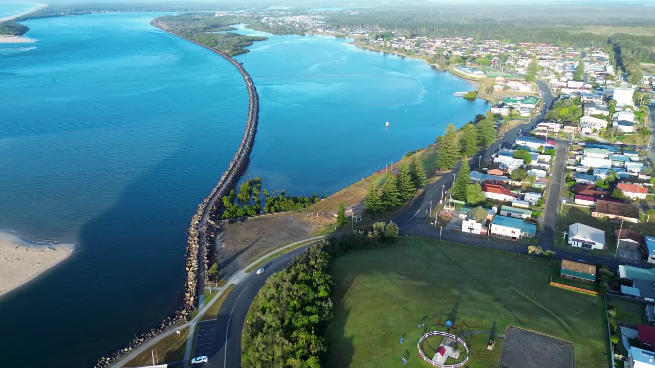 Drone aerial landscape of car driving on road by rural town foreshore waterfront break wall along Manning River with ocean beach dunes and scenic coastline in Harrington NSW Australia travel tourism