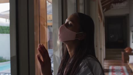 mixed race woman wearing mask at home looking through window