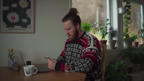 bearded man using mobile phone then laptop while sitting on the wooden table