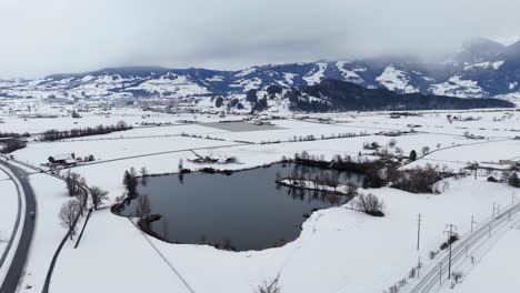 Toma-Aérea-En-órbita-Del-Lago-En-El-Paisaje-Invernal-Nevado-Suizo-Y-Los-Alpes-Como-Telón-De-Fondo