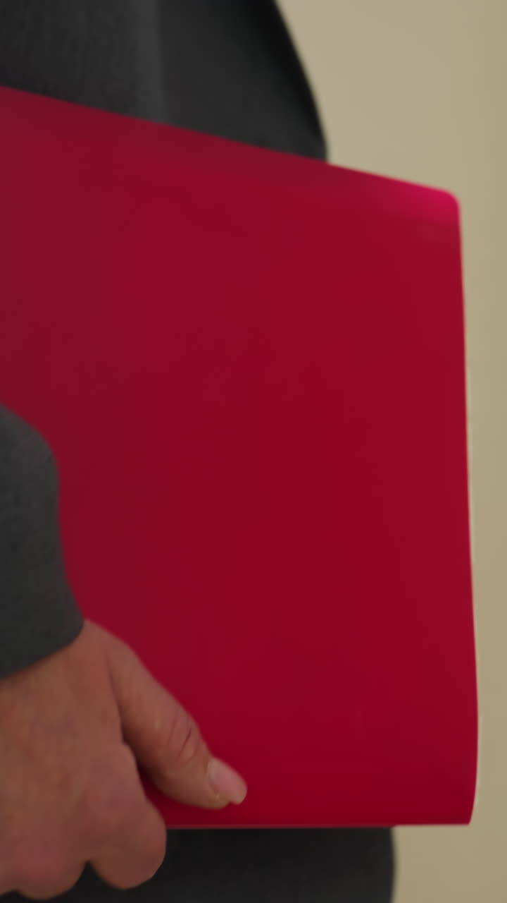 Professional person dressed in gray corporate outfit walking indoors while holding bright red envelope under arm in modern office hallway with sunlight filtering through window in background