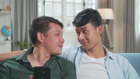 portrait of asian gentle gay couple using mobile phone, while sitting on a couch in cozy stylish apartment. males looking each other