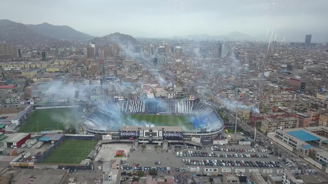 Panoramic aerial view of a packed stadium with fireworks and blue smoke, set against a sprawling cityscape. Ideal for sports, urban, and event imagery.