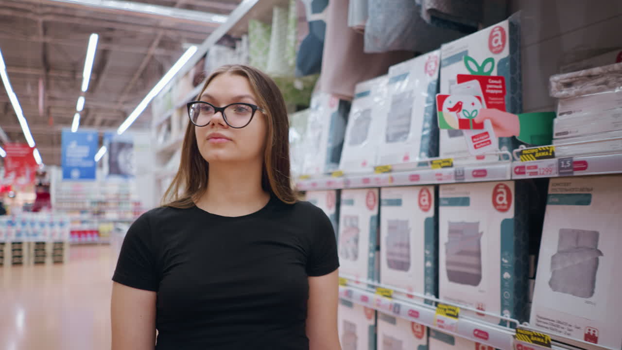 mujer compradora caminando por un gran centro comercial, navegando por los artículos en los estantes mientras mira a su alrededor, mujer elegante con camiseta negra y gafas explorando el espacio minorista con iluminación brillante y productos organizados