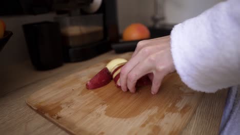 Unrecognizable-woman-at-kitchen-cutting-apple-with-a-knife-on-a-wooden-cutting-board-to-make-juice