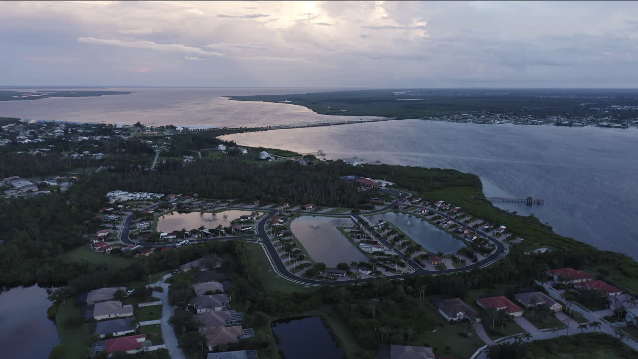 Quiet suburban homes line winding streets beside tranquil ponds and a vast bay, as the soft twilight sky reflects gentle hues of blue and rose across calm evening water