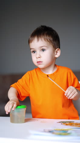 adorable baby boy washing his finger in the cup for brushes. kid with a brush in hand painting with his finger on the paper. vertical video.