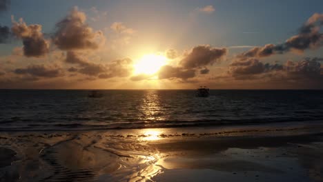 Beautiful-aerial-dolly-in-drone-shot-of-an-ocean-sunrise-at-Well-Beach-near-Joao-Pessoa-on-a-warm-summer-morning-with-the-water-below,-golden-clouds-on-the-skyline,-and-small-boats-in-the-water