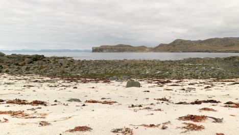 slow panning shot along a sandy beach in scotland looking out over boulders, the ocean and mountains in the background