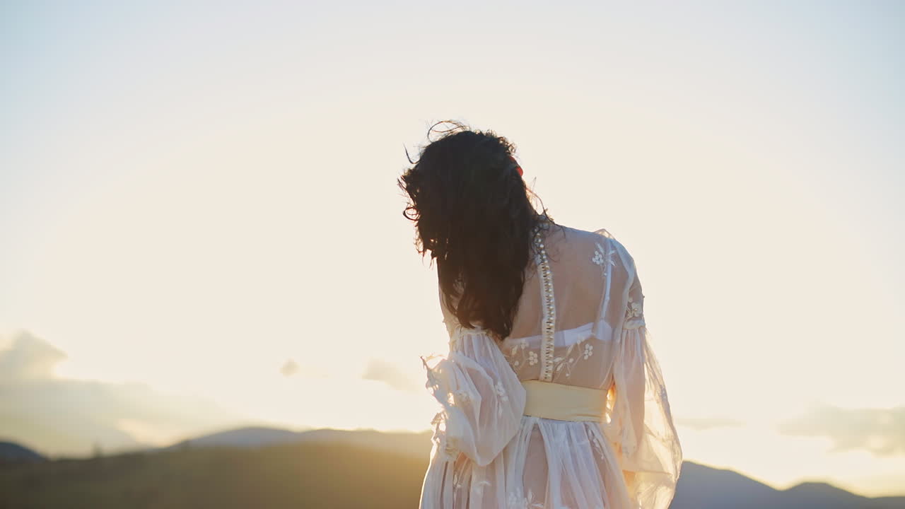 Dark-haired woman in transparent white dress standing high in the mountain. Unrecognizable lady stands her back to the camera enjoying setting sun.