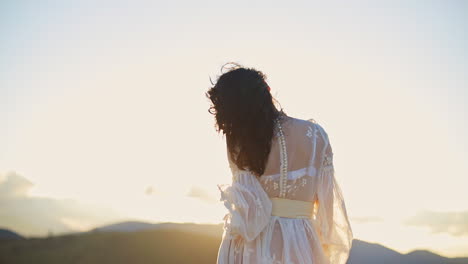 dark-haired woman in transparent white dress standing high in the mountain. unrecognizable lady stands her back to the camera enjoying setting sun.