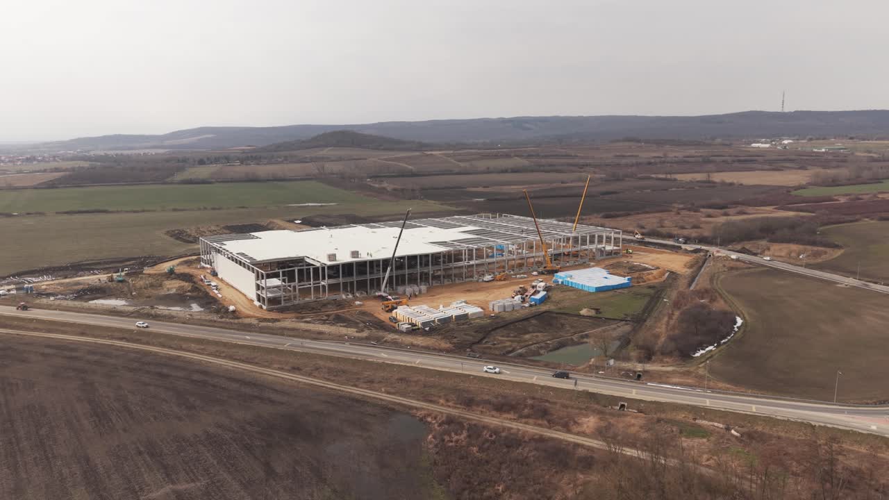 Vista drone di un grande magazzino industriale in costruzione circondato da campi aperti e campagna. Gru, macchinari edili e materiali da costruzione sono visibili intorno alla struttura in acciaio