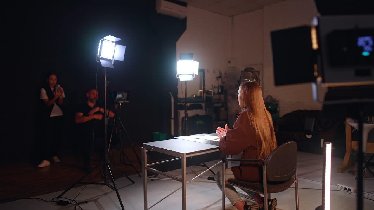 Long-haired brunette woman in brown jacket sits her half back to camera in modern photo studio. Male photographer and female assistant work backstage.