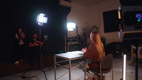 long-haired brunette woman in brown jacket sits her half back to camera in modern photo studio. male photographer and female assistant work backstage.
