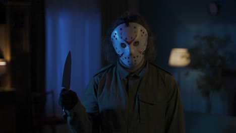 man maniac in white hockey mask standing in the room looking directly at the camera and holding