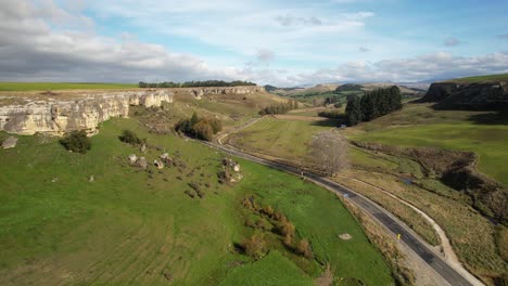 Aerial-of-scenic-road-next-to-limestone-cliff-in-beautiful-valley,-farmland-landscape,-New-Zealand