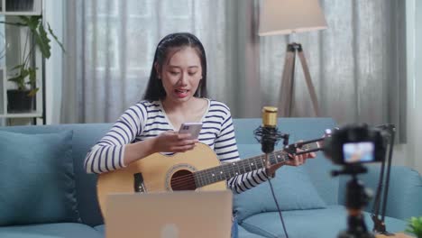 asian woman with a laptop reading comments on smartphone and speaking to camera while live stream playing guitar at home