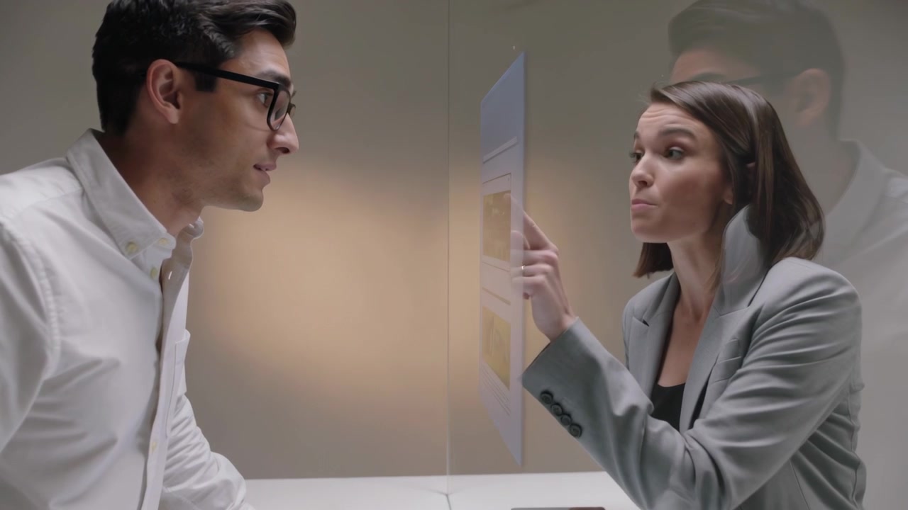 Uomo in camicia bianca che spiega e fa una dimostrazione alla scrivania, con una donna in blazer grigio che risponde dietro una partizione con tablet.
