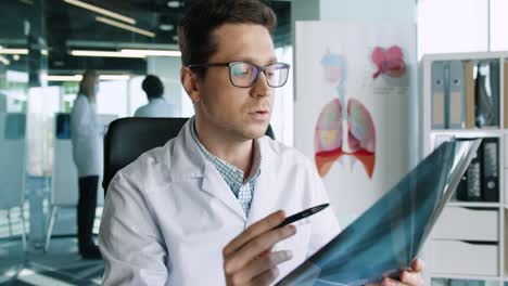 vista de cerca del médico caucásico con anteojos sentado en la clínica en el lugar de trabajo mirando la cámara y explicando la enfermedad pulmonar del coronavirus en la consulta médica